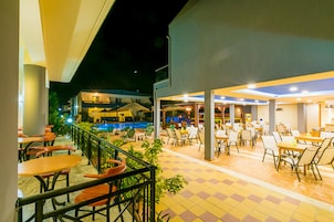 Un área de comedor al aire libre con mesas y sillas, una piscina al fondo y un edificio de varios niveles.