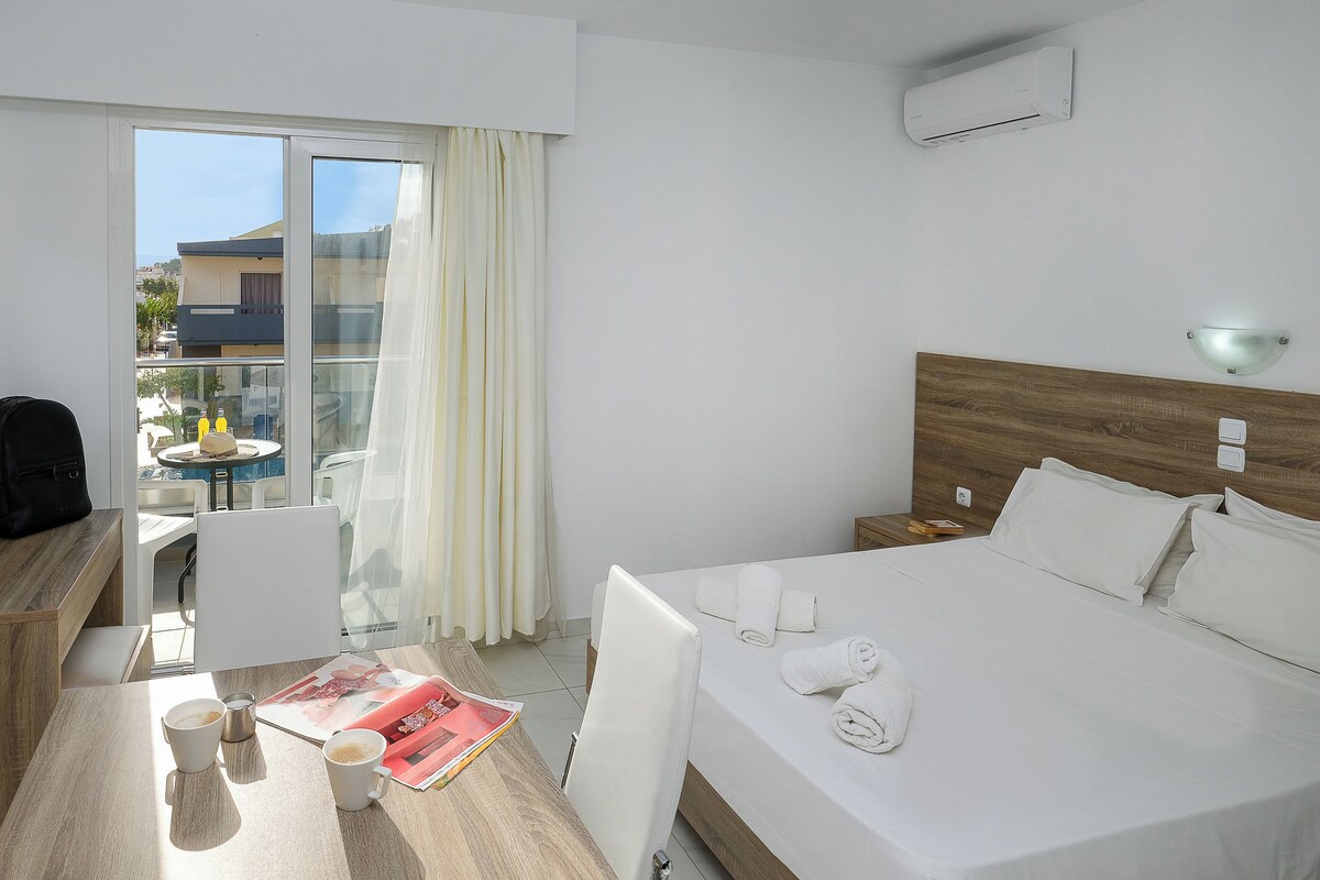 A hotel room with a bed, a desk with a chair, a table with a magazine and coffee cups, and a view of the outside through a sliding door.