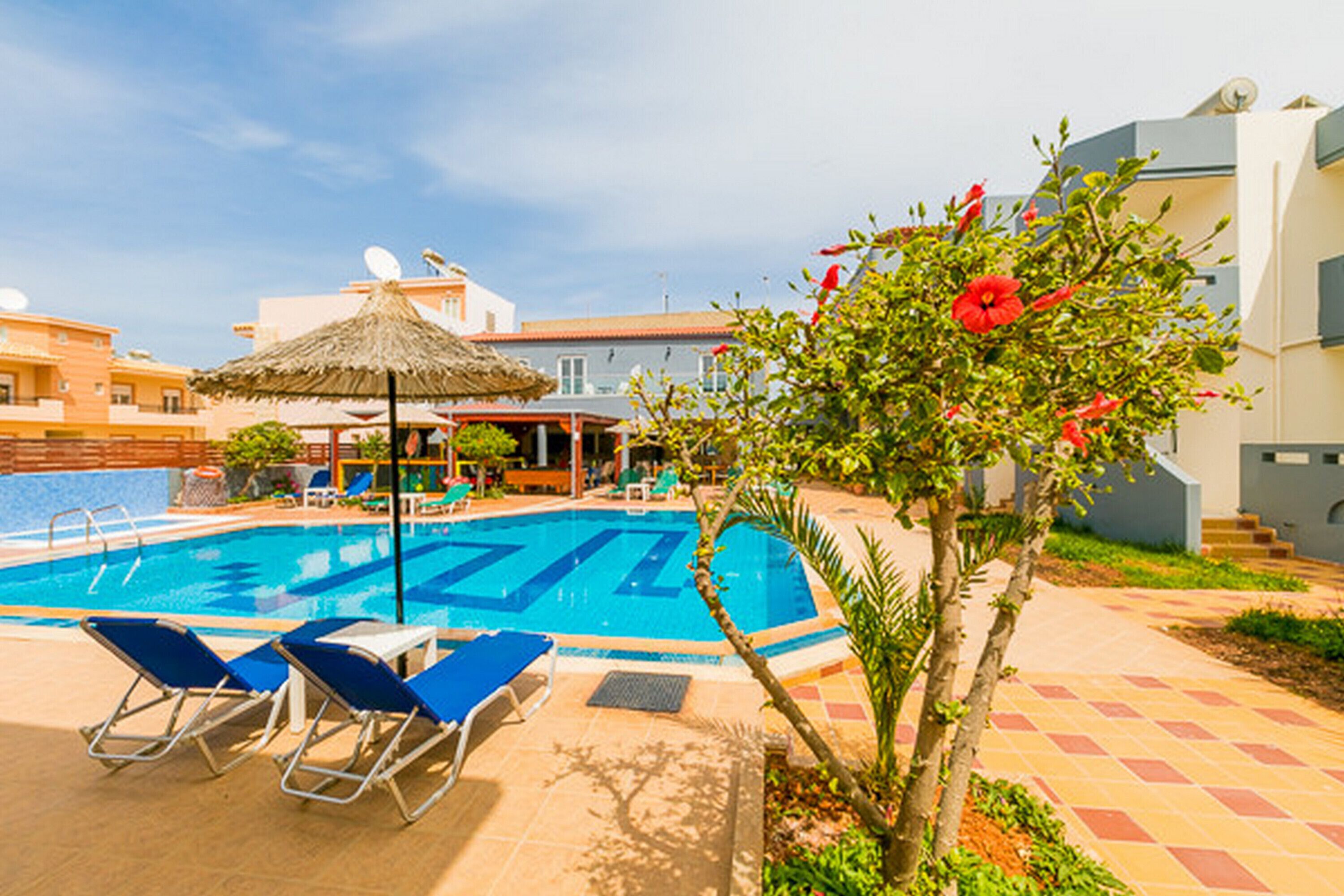 A swimming pool with lounge chairs and a thatched umbrella, surrounded by buildings and greenery.