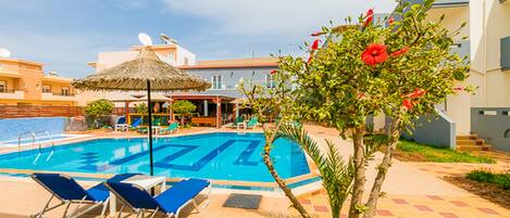 A swimming pool with lounge chairs and a thatched umbrella, surrounded by buildings and greenery.
