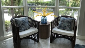 A room with two wicker chairs with patterned cushions, a small round table with a vase of yellow flowers, and large windows offering a view of trees and a street.