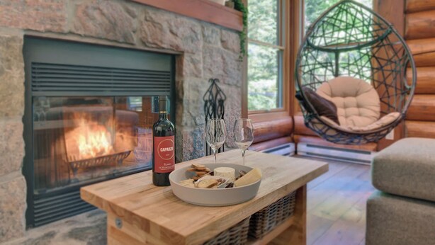 A cozy cabin interior with a stone fireplace, a wooden table with a bowl of snacks, a hanging chair, and a bottle of wine. - Family Lake Access Chalet w/ Spa (Lac-Supérieur)