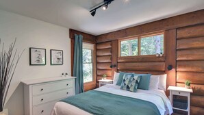 A bedroom with a wooden log wall, a bed with a green blanket, a white dresser, and two bedside tables with plants. - Family Lake Access Chalet w/ Spa (Lac-Supérieur)