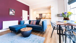 A modern living room with a blue sofa, a wooden coffee table, and a dining area with a table and chairs. - Luxury Apartment w/ Roof Terrace & Parking, Town Centre (Great Yarmouth)
