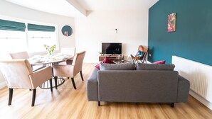A modern living room with a grey sofa, dining table with chairs, a TV, and artwork on teal walls.