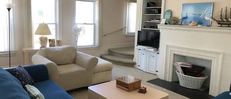 A living room with a beige sofa, blue armchairs, a coffee table, a fireplace, a TV, and a staircase leading to another room.