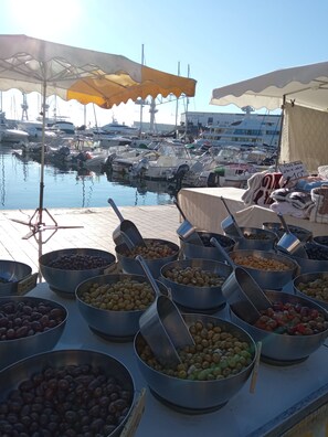 Een jachthaven met aangemeerde boten, een kraam met verschillende soorten fruit en olijven, en een restaurant op de achtergrond.