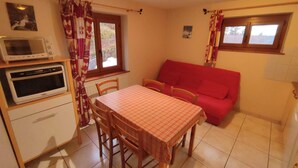 Cocina compacta con microondas, horno y mesa de comedor con sillas. Hay un sofá rojo y una ventana con cortinas.
