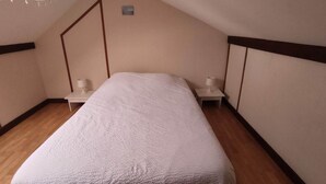 Una cama individual en una habitación con suelo de madera, paredes blancas y techo inclinado. Hay dos mesitas de noche con lámparas.