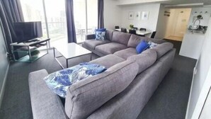 A modern living room with a grey sectional sofa, a glass coffee table, and a flat-screen TV. - Chevron Renaissance, 2BR w/ Views, Parking & WIFI (Gold Coast)