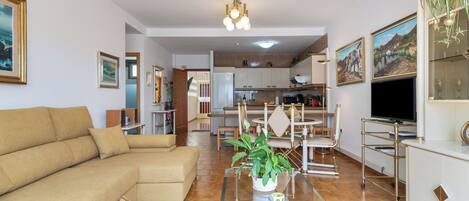 A living room with a sofa, a glass coffee table with a plant, a kitchen in the background, and a TV on a stand.