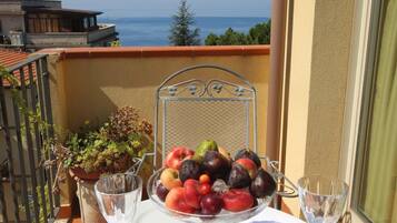 Une table dressée pour deux personnes, avec un panier de fruits, des verres à vin et des couverts, sur un balcon donnant sur la mer.