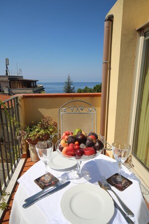 Une table dressée pour deux personnes, avec un panier de fruits, des verres à vin et des couverts, sur un balcon donnant sur la mer.