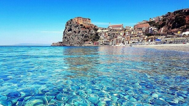 Un pueblo costero con aguas azules cristalinas, un acantilado rocoso imponente y edificaciones en la ladera.