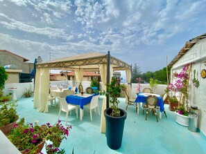 Un patio exterior con mesas y sillas, una pérgola, plantas en macetas y vista al mar.