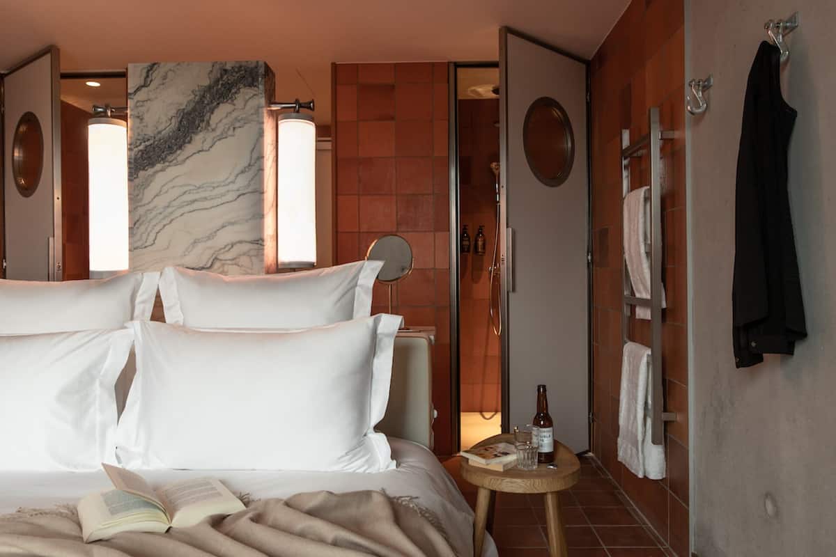 Una habitación de hotel con cama, un espejo redondo, una mesita con una botella y una zona de ducha.