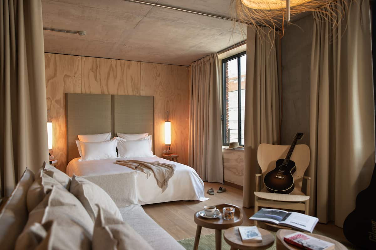 Habitación de hotel con una cama grande, una silla con una guitarra, una mesita con un libro y una taza, y una ventana con cortinas.