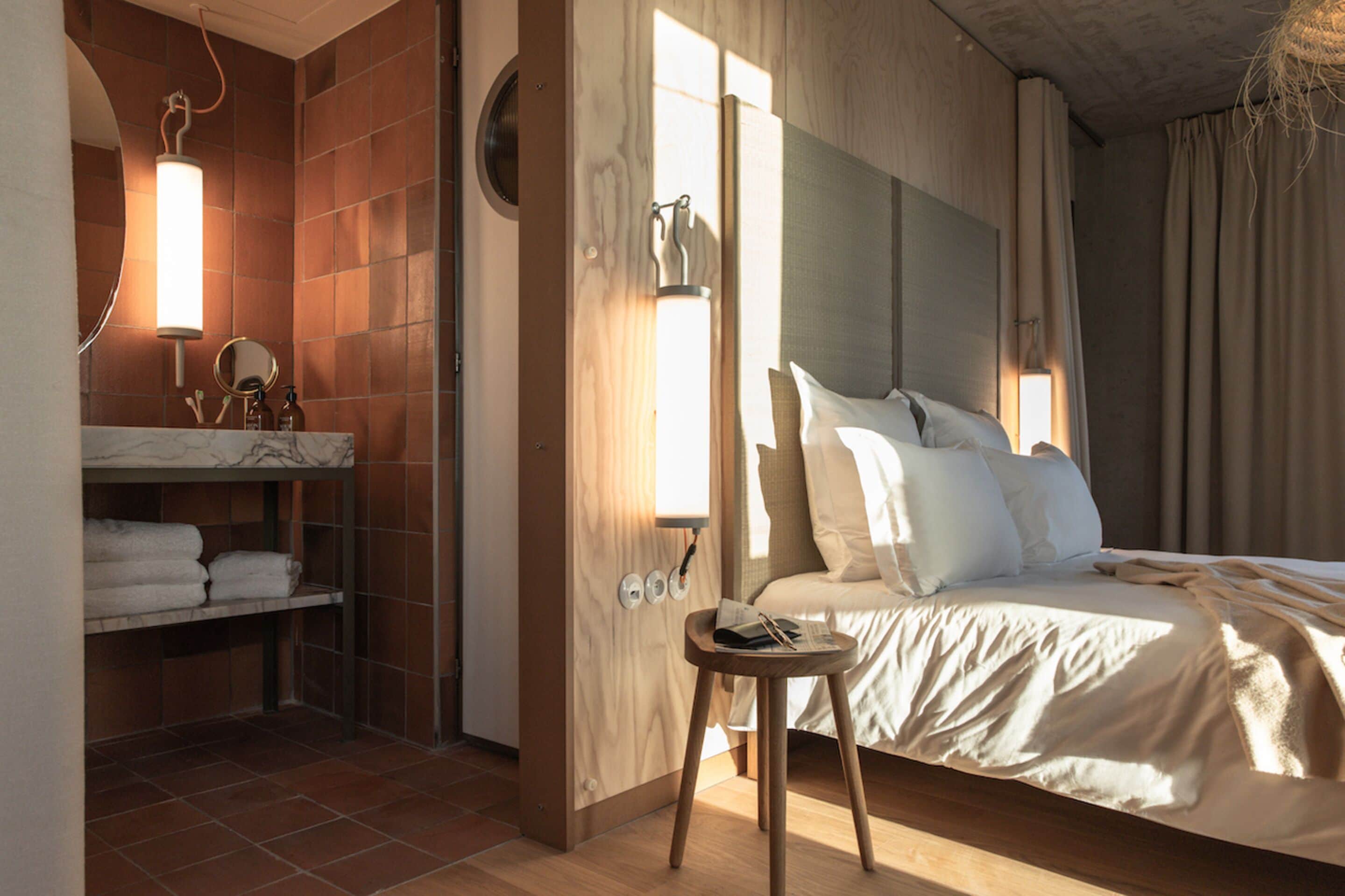A hotel room with a bed, a bedside table, a mirror, a shelf with towels, and a window with curtains.
