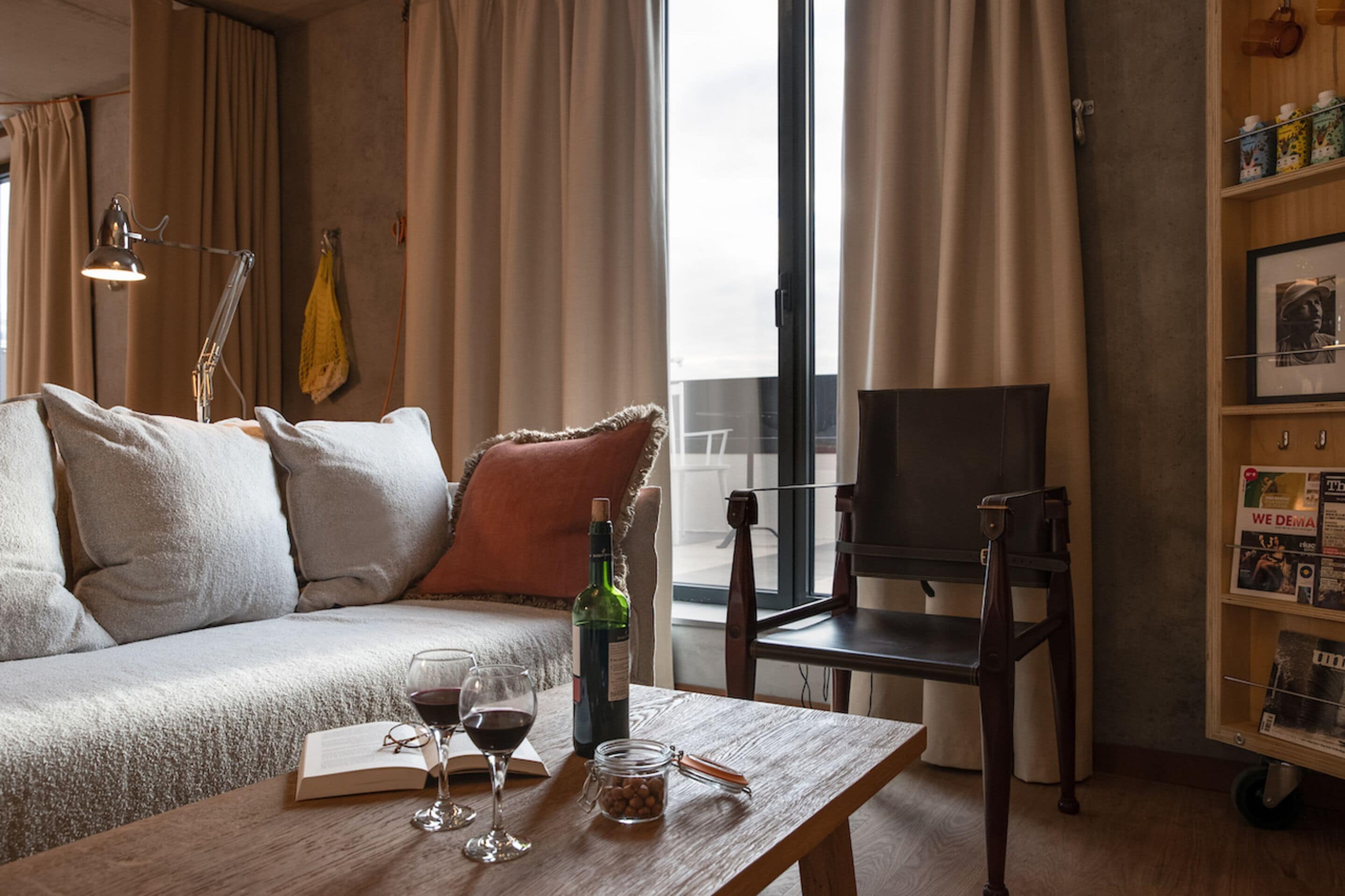 Una sala de estar acogedora con un sofá, una mesa de centro con un libro, copas de vino y una botella. Hay un televisor en un mueble bajo y una estantería con varios objetos.