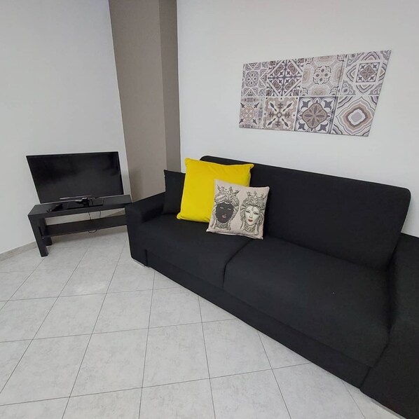 A living room with a black sofa, a TV on a stand, and a patterned wall hanging. - Focu e Mari (Mascali)