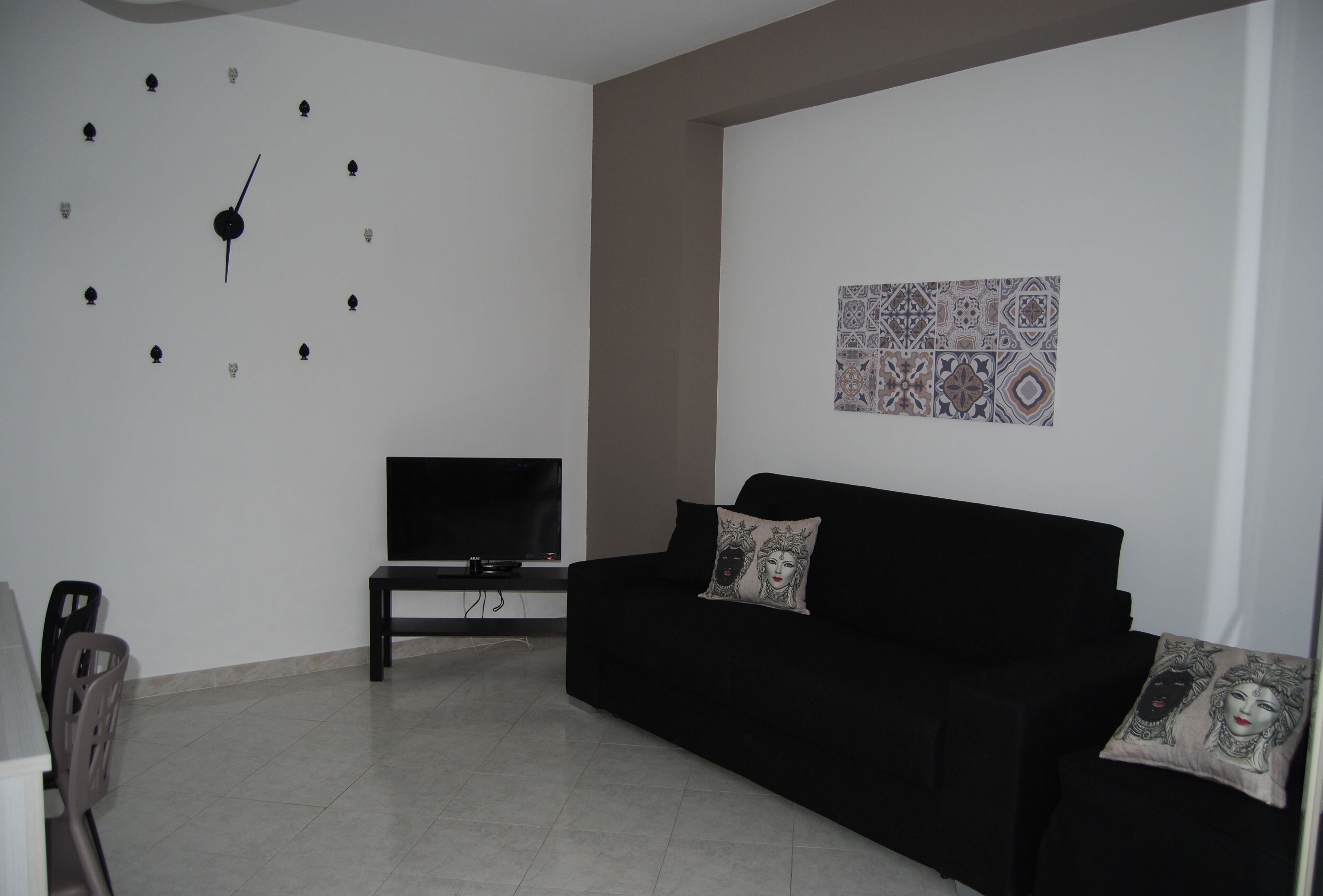 Un salon avec un canapé noir, un téléviseur à écran plat posé sur un meuble, une horloge murale et un élément de décoration murale à motifs.