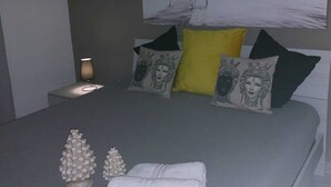A modern bedroom with a grey bedspread, two decorative pillows, a yellow pillow, a nightstand with a lamp, and a wall-mounted artwork. - Focu e Mari (Mascali)
