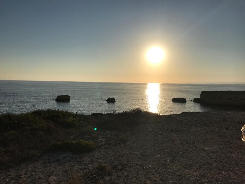 A coastal sunset with the sun low on the horizon, calm sea, and rocky outcrops.