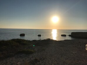 A coastal sunset with the sun low on the horizon, calm sea, and rocky outcrops. - Focu e Mari (Mascali)