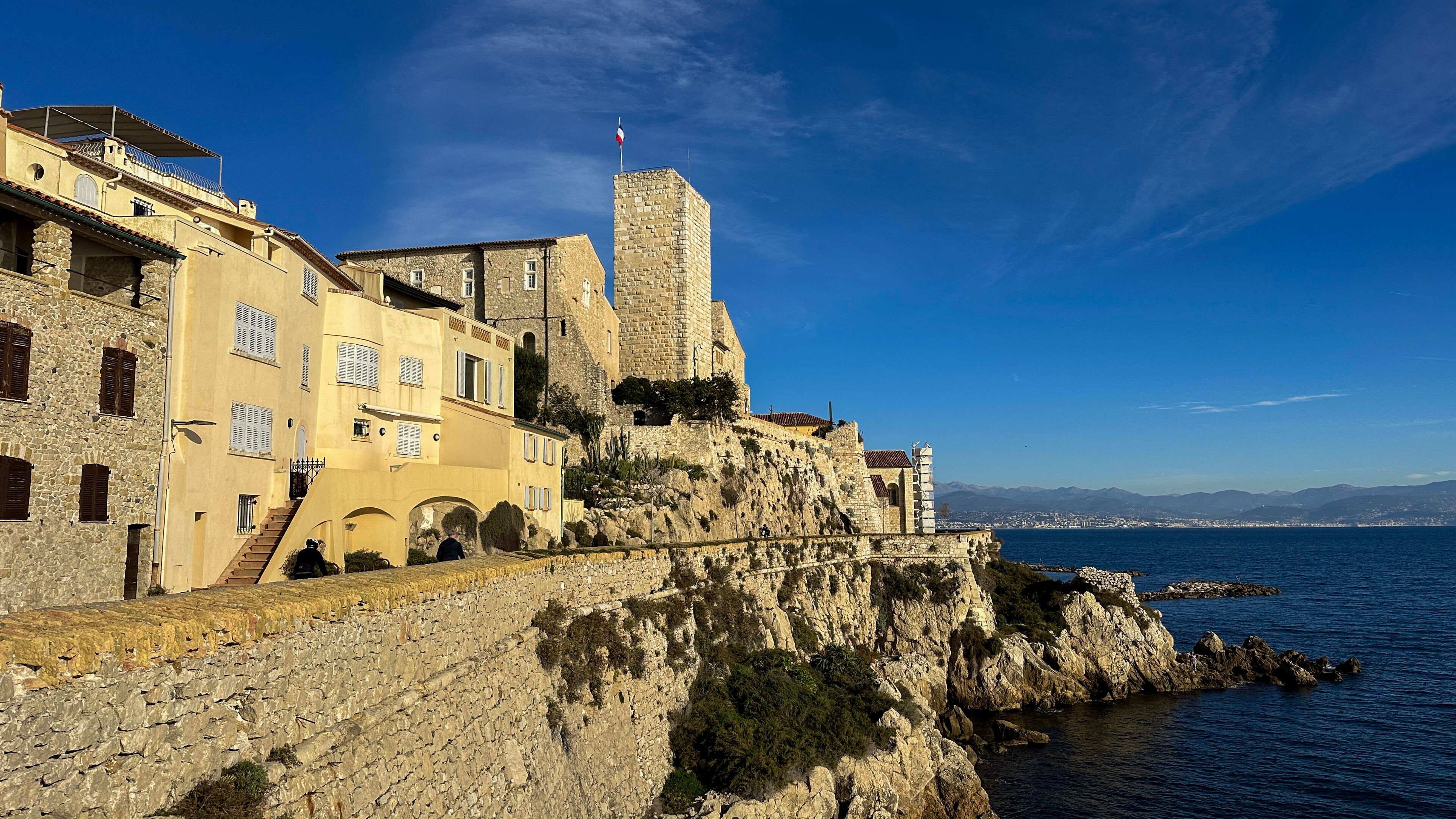 A coastal town with historic stone buildings, a prominent tower, and a view of the sea.