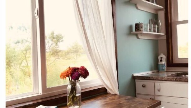 A kitchen with a wooden table, a chair, a vase with flowers, and a window with curtains. - Grinnell Heritage Tiny House - On Organic Farm (Grinnell)