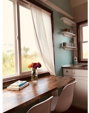 A kitchen with a wooden table, a chair, a vase with flowers, and a window with curtains. - Grinnell Heritage Tiny House - On Organic Farm (Grinnell)
