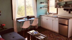 A compact kitchen with white cabinets, a built-in microwave, and a sink. There is a window with sheer curtains, a small table with chairs, and a colorful rug on the floor. - Grinnell Heritage Tiny House - On Organic Farm (Grinnell)