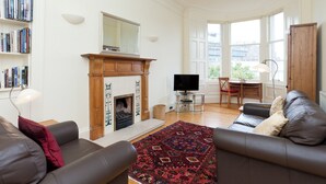 Ein Wohnzimmer mit Kamin, Fernseher, einer Couch, einem Bücherregal und großen Fenstern.