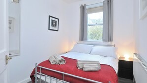 Un dormitorio con cama, ventana con cortinas, mesita de noche con lámpara y un cuadro enmarcado en la pared.