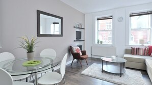 Una sala de estar moderna con una mesa de comedor de cristal, sillas blancas, un sillón marrón, un sofá beige y una alfombra blanca.