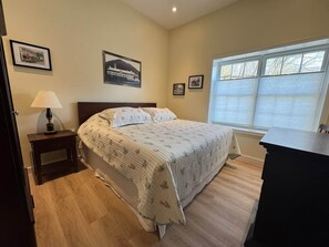 A bedroom with a bed, a nightstand, a lamp, a window with blinds, and framed pictures on the wall.