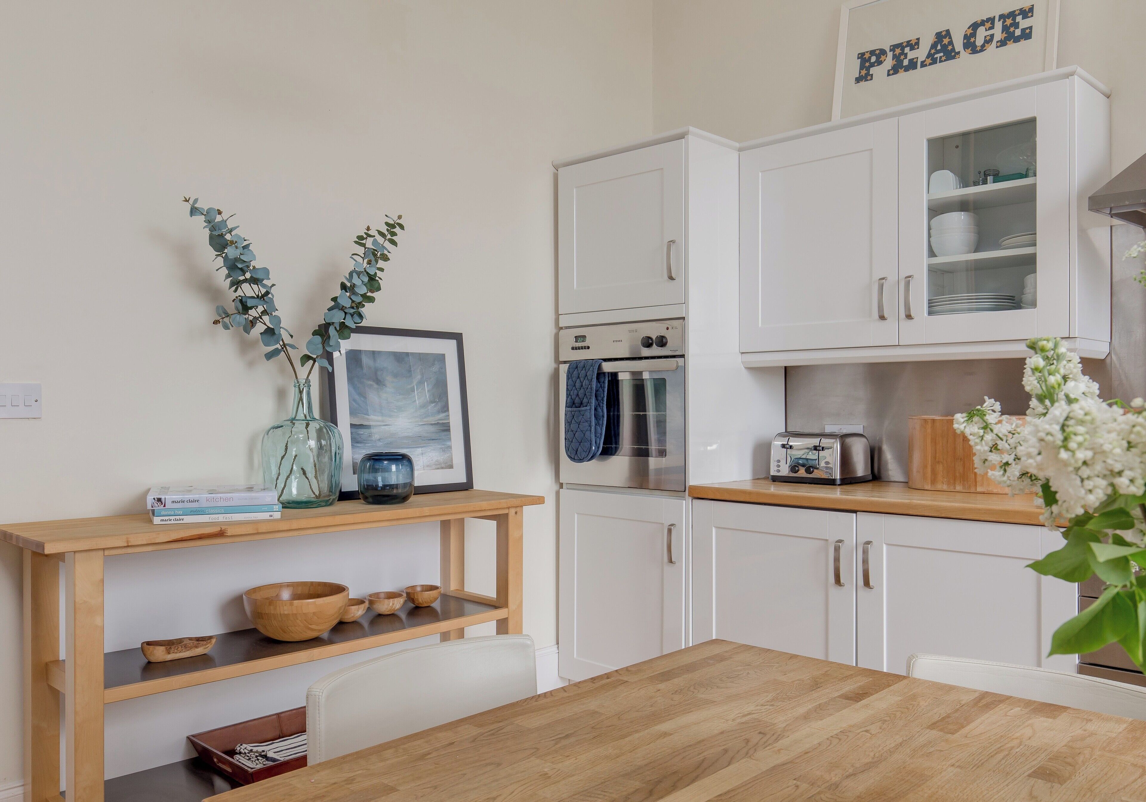 Une cuisine moderne avec des meubles blancs, une table en bois et un plan de travail sur lequel on trouve un grille-pain et un vase de fleurs.