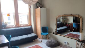 Una sala de estar con un sofá gris, un sillón azul, un otoman redondo, un tapete estampado y un espejo grande.