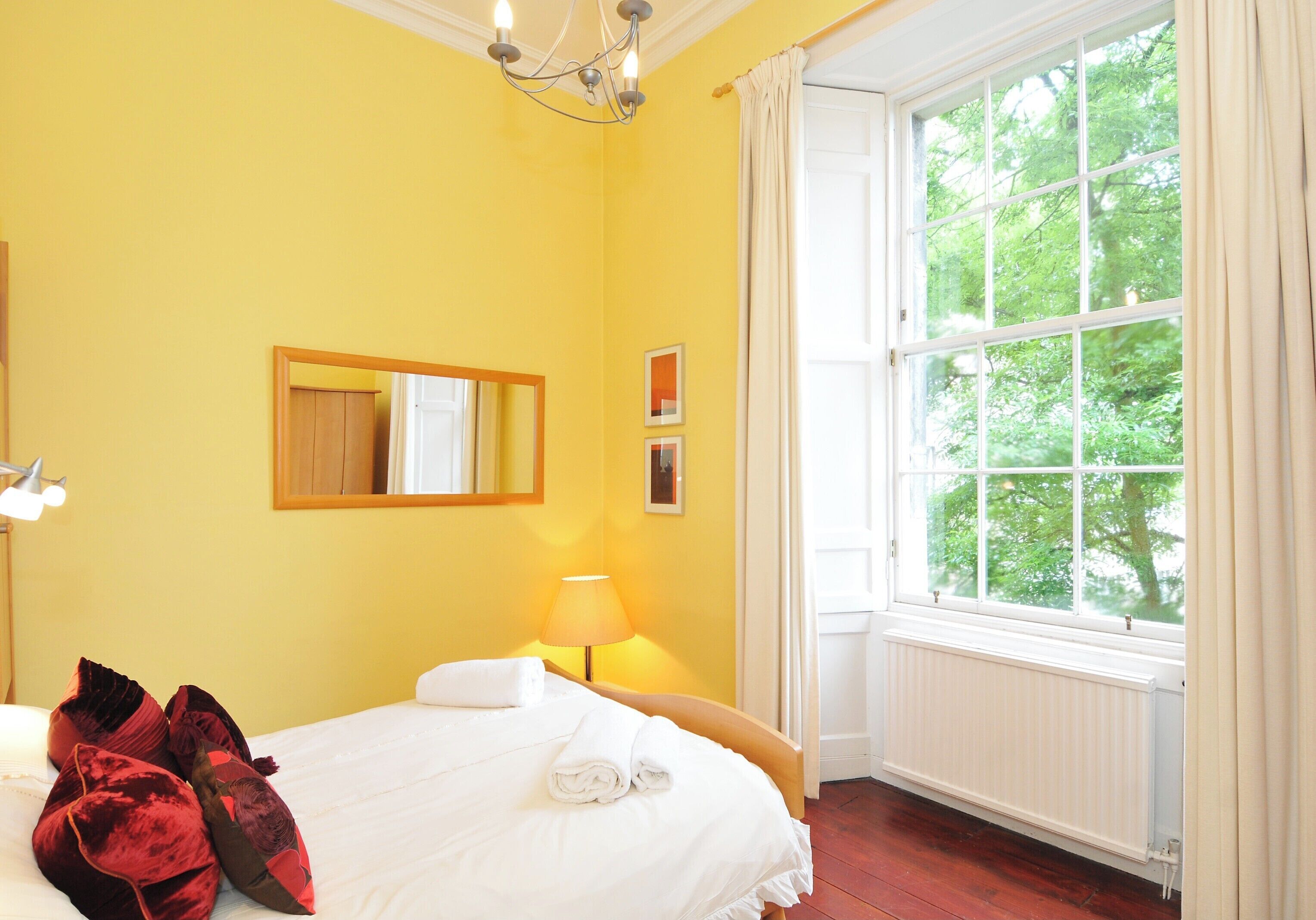 A hotel room with a large window, a bed with white linens, a lamp, and a mirror on the wall.