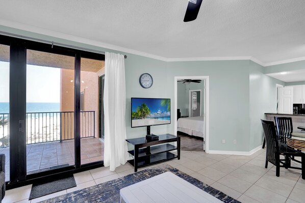 A living room with a TV, a dining area, and a balcony with a view of the beach.