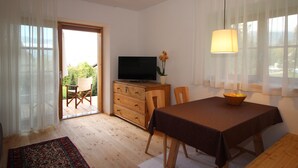 A room with a wooden floor, a TV on a wooden cabinet, a dining table with a brown tablecloth, and a chair.