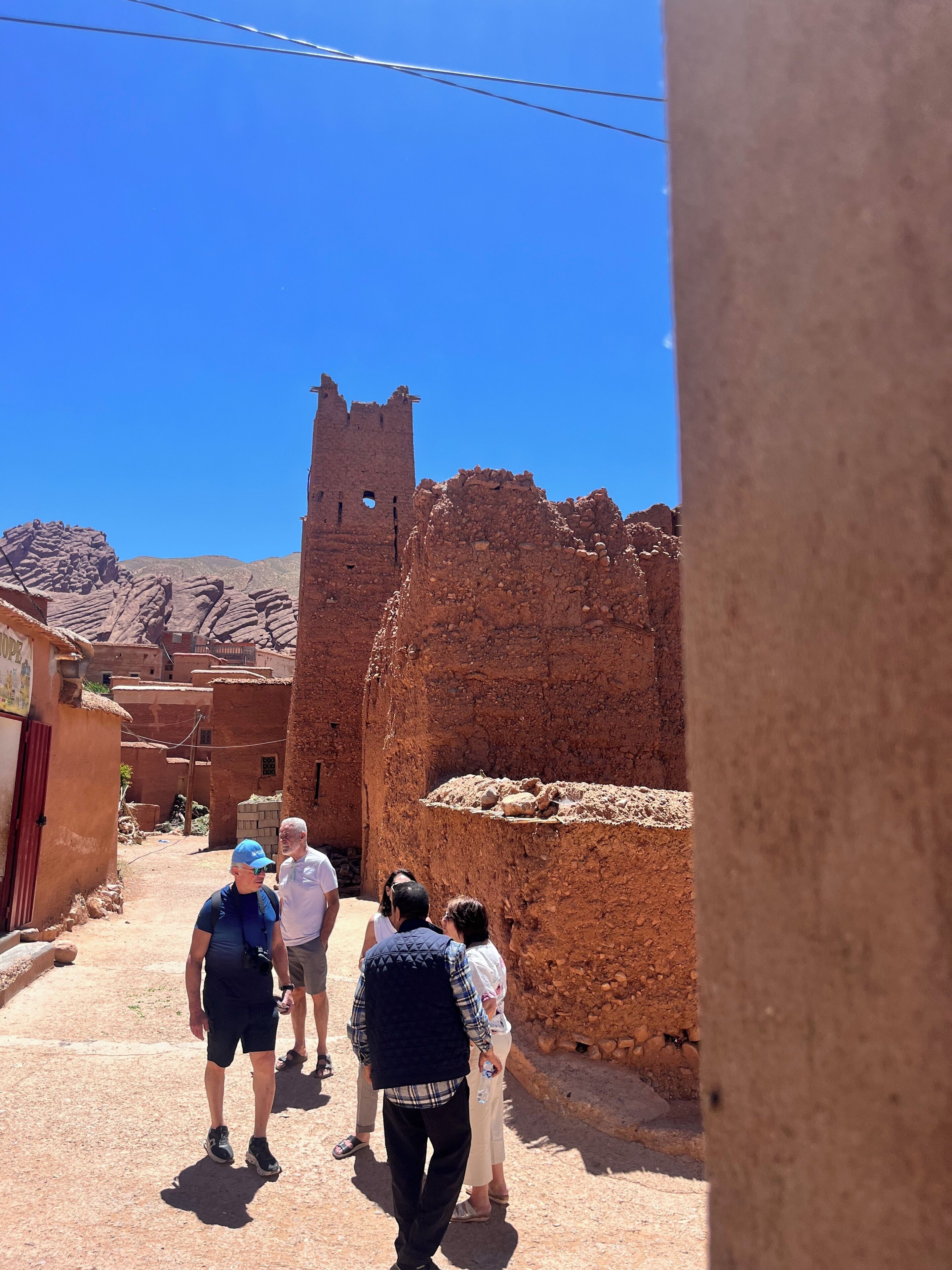 A group of people walking past a historic stone structure with a tower, in a narrow alleyway with traditional buildings.