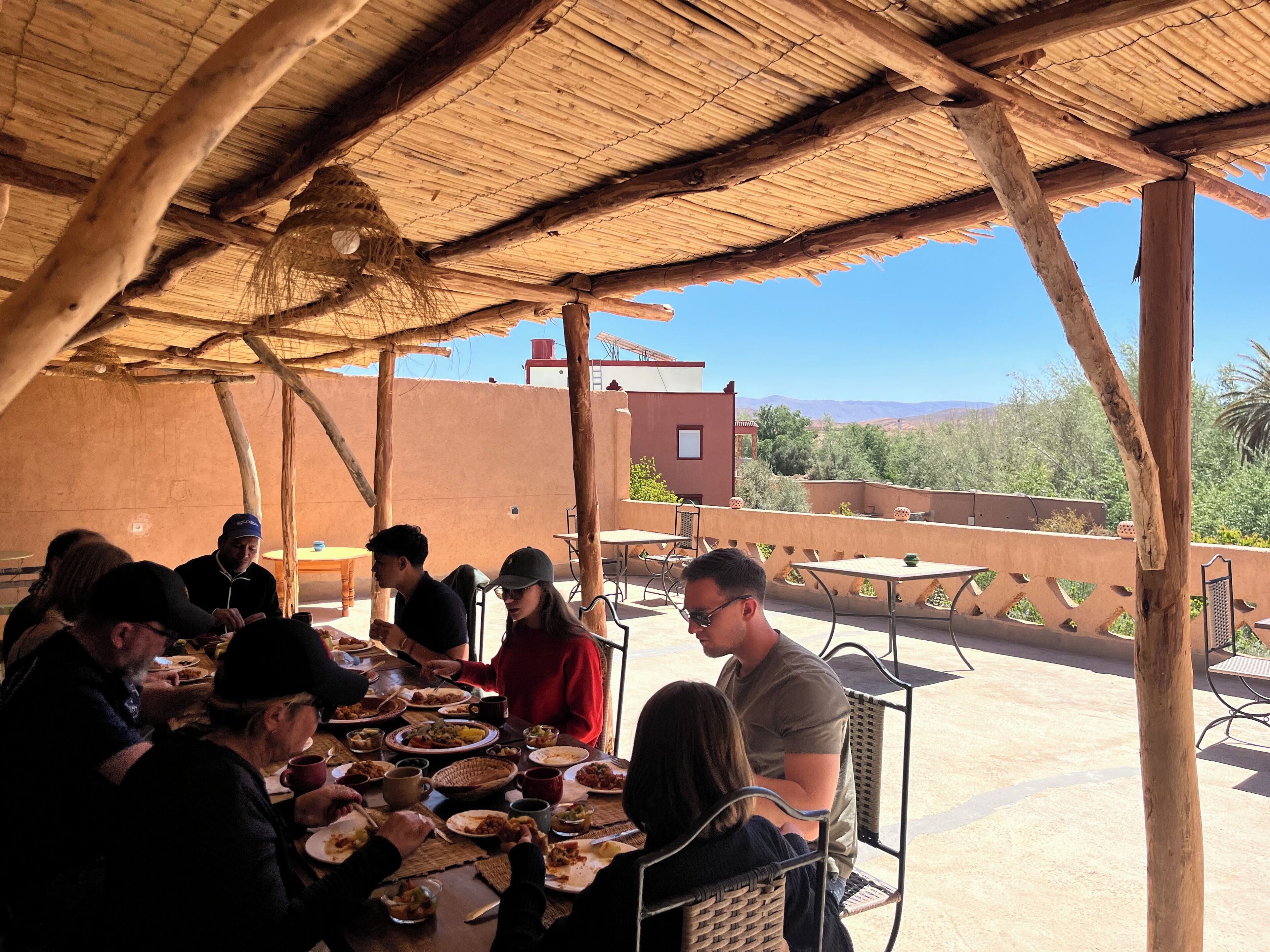Un groupe de personnes est assis à une table en plein air, sous une structure en bois, et profite d’une vue sur un bâtiment et un paysage en arrière-plan.