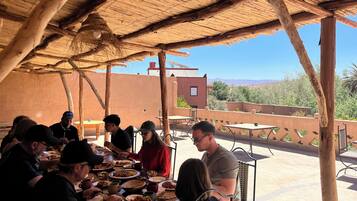Un groupe de personnes est assis à une table en plein air, sous une structure en bois, et profite d’une vue sur un bâtiment et un paysage en arrière-plan.