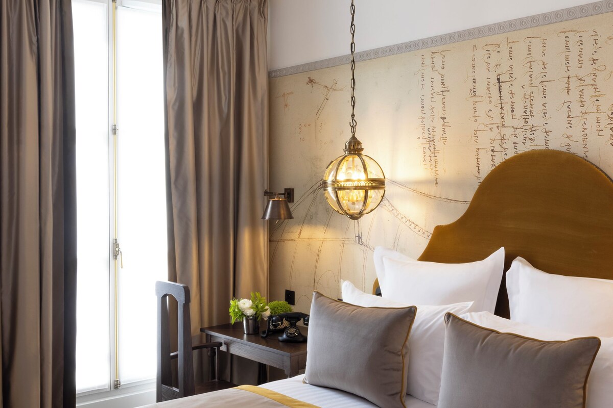 Una habitación de hotel con una cama grande, un cabecero de madera, una mesita de noche con lámpara y vistas al exterior a través de una ventana con cortinas.