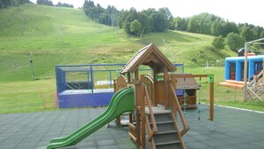 Une aire de jeux avec un toboggan vert, des structures d’escalade en bois et une zone délimitée par une clôture.