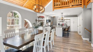 Ein geräumiger Essbereich mit einem langen Holztisch und weißen Stühlen, eine Küche mit weißen Schränken und eine Holztreppe, die in eine obere Etage führt.