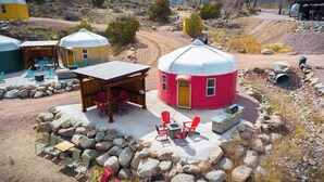 Una yurta con un área de estar al aire libre techada, rodeada de rocas y un sendero de tierra.