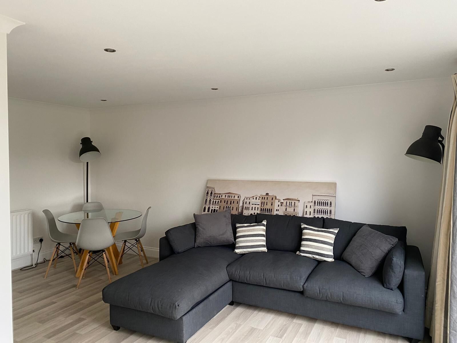 A modern living room with a grey sectional sofa, a glass dining table with chairs, and a cityscape artwork on the wall.