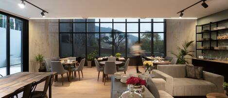 A modern living room with a wooden dining table, grey sofas, and a large window offering a view of trees and mountains.